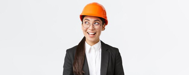 Primer plano de una arquitecta asiática creativa que tiene una idea interesante en mente ingeniero de construcción en traje de negocios y casco de seguridad mirando a la izquierda con una sonrisa complacida de pie fondo blanco