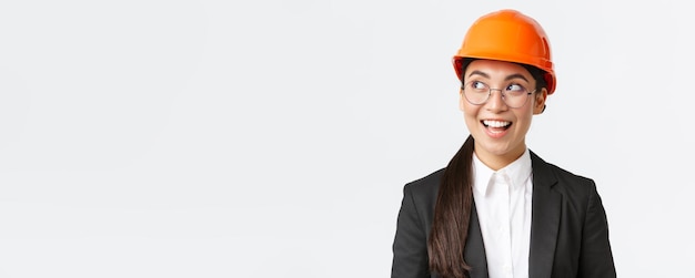 Primer plano de una arquitecta asiática creativa que tiene una idea interesante en mente ingeniero de construcción en traje de negocios y casco de seguridad mirando a la izquierda con una sonrisa complacida de pie fondo blanco