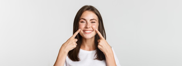 Primer plano de una atractiva joven sonriente señalando con el dedo la cara metiendo las mejillas en el fondo blanco