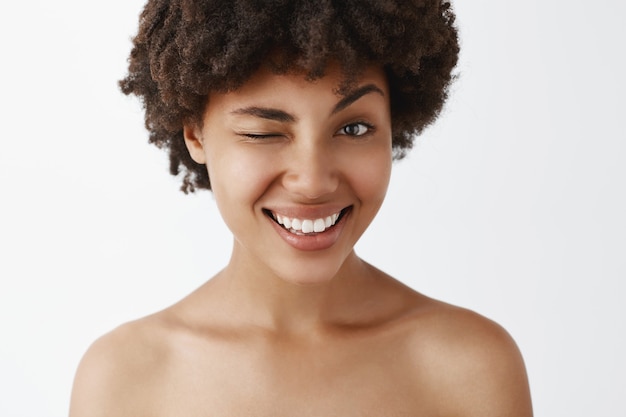 Primer plano de una atractiva modelo de piel oscura, emotiva, feliz y de aspecto amistoso con peinado afro posando desnuda, sonriendo ampliamente y guiñando un ojo como si insinuara algún concepto o secreto interesante