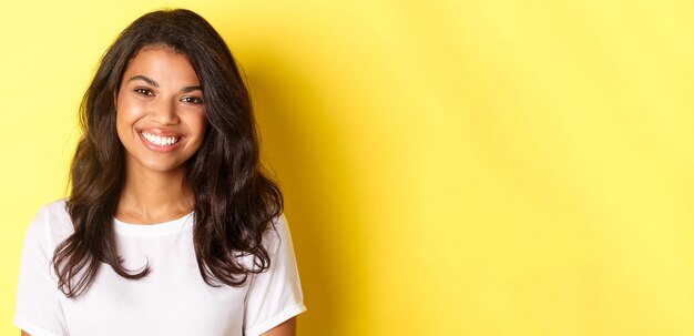 Primer plano de una atractiva mujer afroamericana sonriendo y luciendo feliz de pie sobre fondo amarillo