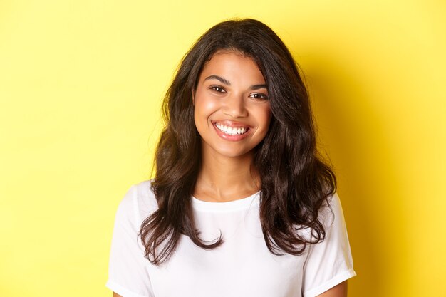 Primer plano de una atractiva mujer afroamericana, sonriendo y mirando feliz, de pie sobre fondo amarillo