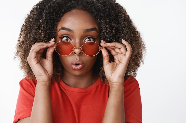 Primer plano de una atractiva mujer afroamericana sorprendida y divertida con el pelo rizado quitándose las gafas de sol y doblando los labios de asombro e interés reaccionando a la impresionante escena