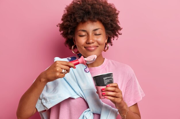 Primer plano de una atractiva mujer de pelo rizado se encuentra con los ojos cerrados, disfruta del agradable sabor del helado de fresa, viste un suéter sobre el hombro, aislado en la pared rosa. Delicioso postre frío