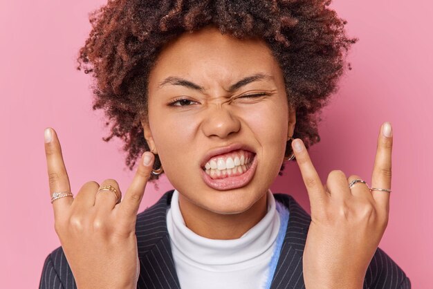 Primer plano de una atrevida mujer de pelo rizado segura de sí misma que hace gestos de signos de rock n roll disfruta activamente de los fruncimientos de ceño de heavy metal meciéndose en el interior aislado sobre un fondo rosa. Concepto de lenguaje corporal