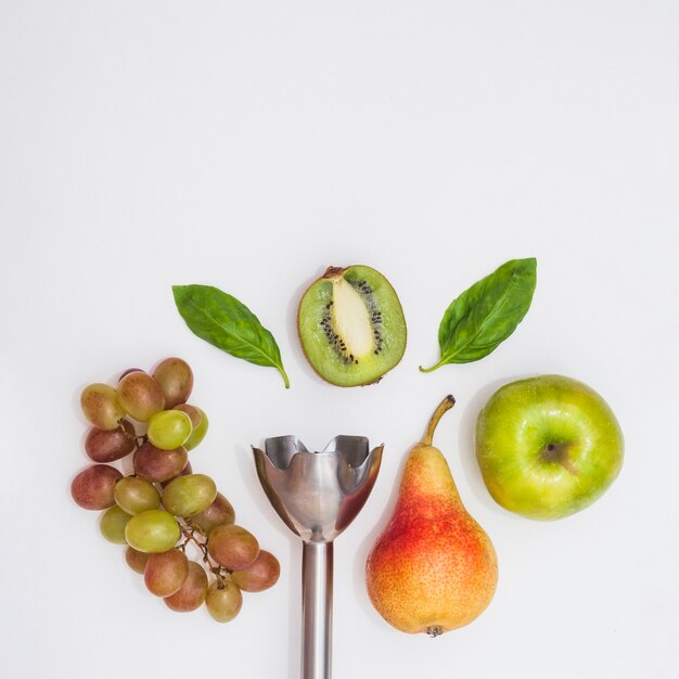 Primer plano de la batidora eléctrica de mano con uvas; peras; manzana; kiwi a la mitad y albahaca sobre fondo blanco