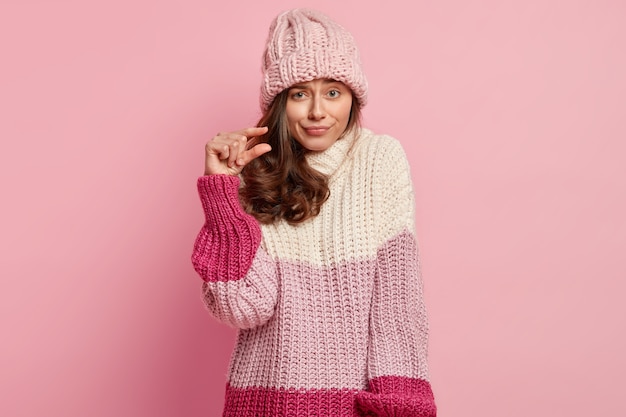 Primer plano de una bella dama forma algo diminuto, hace un gesto con la mano, tiene una expresión facial disgustada, viste ropa de invierno de moda, posa contra la pared rosa. Muy pequeño o pequeño