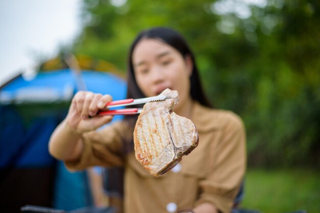 Primer plano de bistec de cerdo feliz joven mujer asiática sosteniendo bistec de cerdo con pinzas mientras se sienta en una silla en el camping