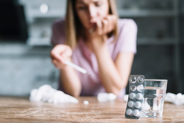 Primer plano de blister y vaso de agua con una mujer enferma en el fondo