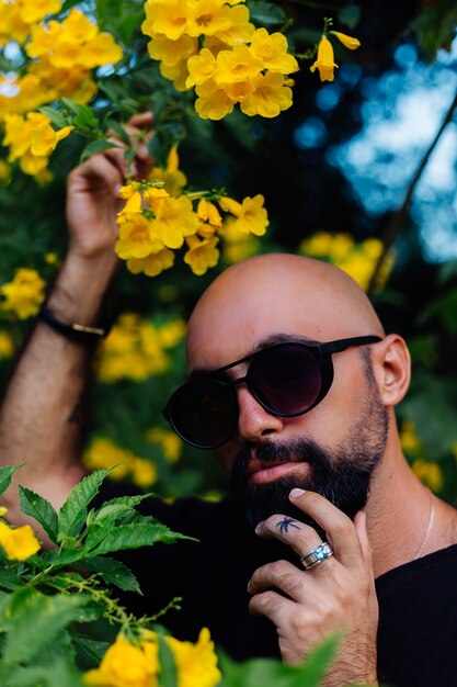 Primer plano de brutal hombre barbudo bronceado con gafas de sol con tatuaje de palmera en los soportes de los dedos rodeado de flores amarillas en el parque