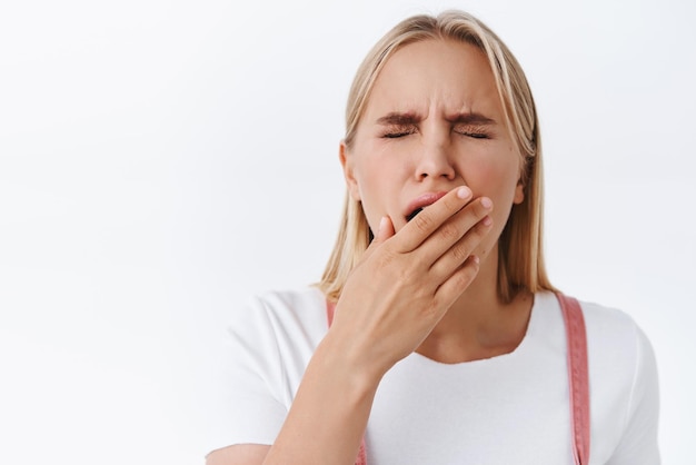 Primer plano cansado bostezando linda mujer rubia en la cubierta de la camiseta abrió la boca con la palma mientras se quedaba dormido sintiendo fatiga o exhausto cerrar los ojos despertar temprano en la mañana quiero café fondo blanco