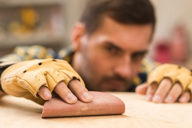 Primer plano de un carpintero macho con guantes de protección en la mano frotando papel de lija en tablón de madera