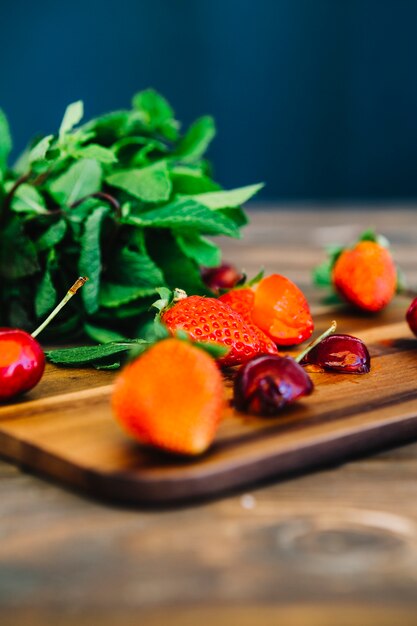 Primer plano de cerezas y fresas en tabla de cortar frente a hojas de menta