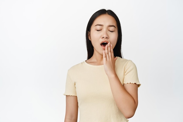 Primer plano de una chica asiática bostezando con los ojos cerrados sintiendo sueño de pie en camiseta amarilla sobre fondo blanco