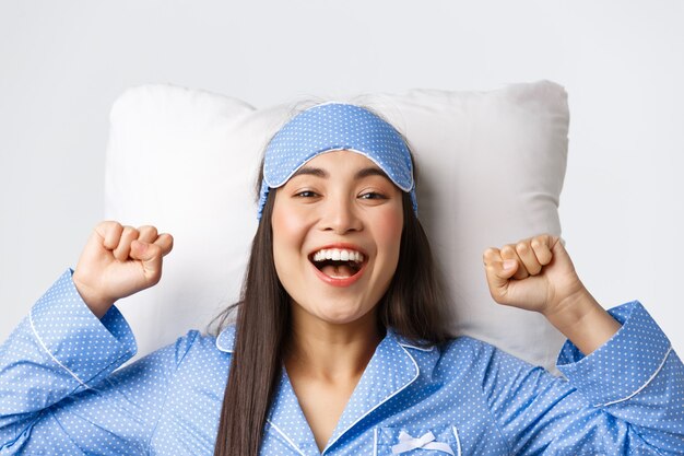 Primer plano de una chica asiática entusiasta en pijama azul y antifaz para dormir, estirando las manos hacia arriba encantados después de dormir bien, quitarse la máscara por la mañana, acostada en la cama sobre la almohada y sonriendo feliz