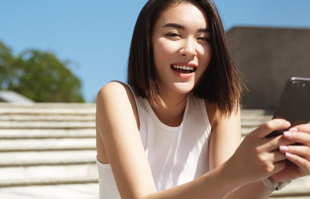 Primer plano de una chica asiática moderna sentada en las escaleras de la calle y sosteniendo un teléfono inteligente Mujer joven descansando al aire libre enviando un mensaje de texto de navegación por Internet