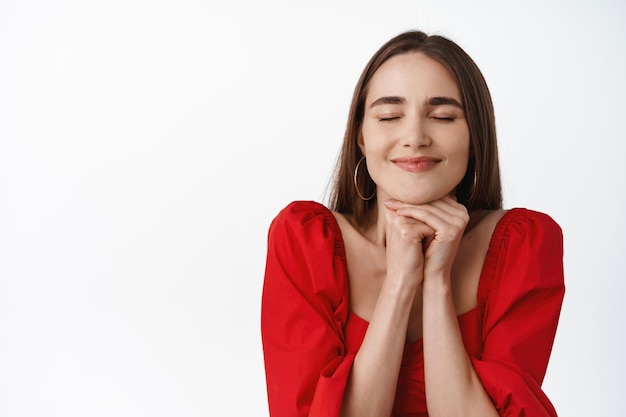 Primer plano de una chica soñadora feliz pensando, cerrar los ojos y parecer esperanzada, imaginar algo, usar un vestido rojo y soñar despierto, desear que suceda algo, fondo blanco