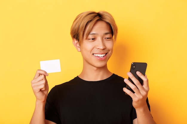 Primer plano de un chico asiático con estilo guapo de compras en línea, mirando el teléfono móvil y sonriendo, mostrando la tarjeta de crédito, de pie sobre la pared amarilla.
