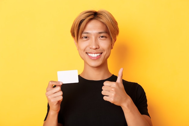 Foto gratuita primer plano de chico asiático satisfecho recomendar banco, mostrando tarjeta de crédito y de pie sobre una pared amarilla
