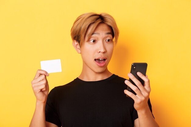 Primer plano de un chico asiático sorprendido mirando el teléfono móvil mientras compra en línea y muestra la tarjeta de crédito, pared amarilla
