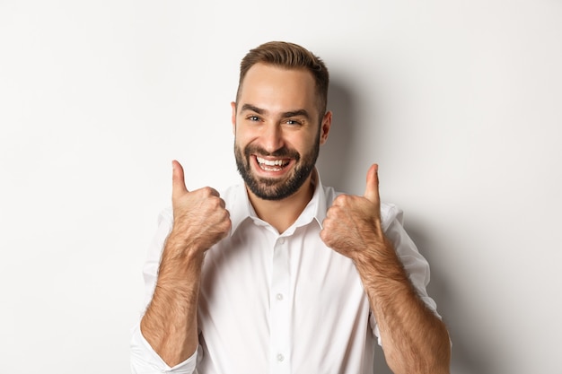 Foto gratuita primer plano de chico barbudo satisfecho con camisa blanca, mostrando los pulgares hacia arriba en señal de aprobación, como y de acuerdo, respuesta positiva