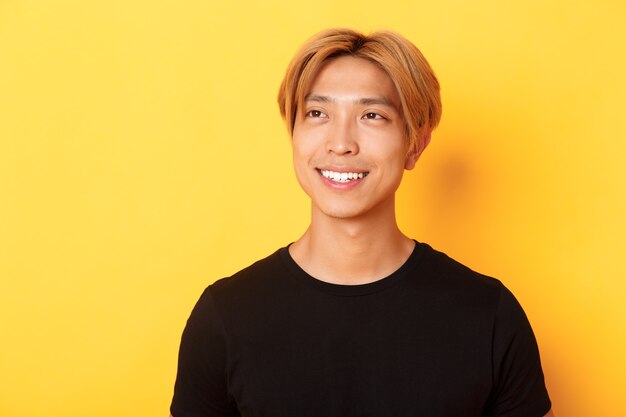 Primer plano de un chico coreano elegante y guapo con cabello rubio, con aspecto soñador y esperanzado en la esquina superior izquierda, sonriendo complacido, de pie en la pared amarilla