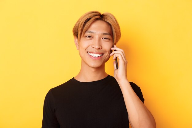 Primer plano de chico coreano con estilo guapo sonriendo y hablando por teléfono móvil, de pie sobre la pared amarilla