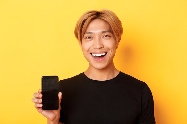 Primer plano de un chico coreano guapo con estilo mostrando la pantalla del teléfono inteligente y sonriendo complacido, recomendar la aplicación móvil, de pie sobre la pared amarilla