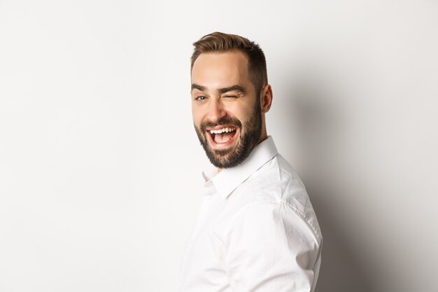 Primer plano de chico descarado con barba, gira la cara a la cámara y guiña un ojo con una sonrisa, de pie