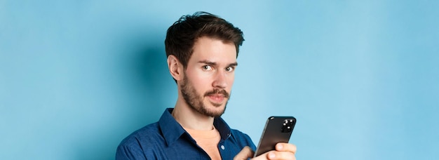 Primer plano de un chico europeo guapo usando un teléfono inteligente y mirando a la cámara de pie sobre fondo azul