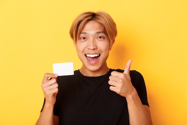 Primer plano de un chico guapo asiático feliz y satisfecho, mostrando la tarjeta de crédito y el pulgar hacia arriba en señal de aprobación, sonriendo asombrado, de pie pared amarilla