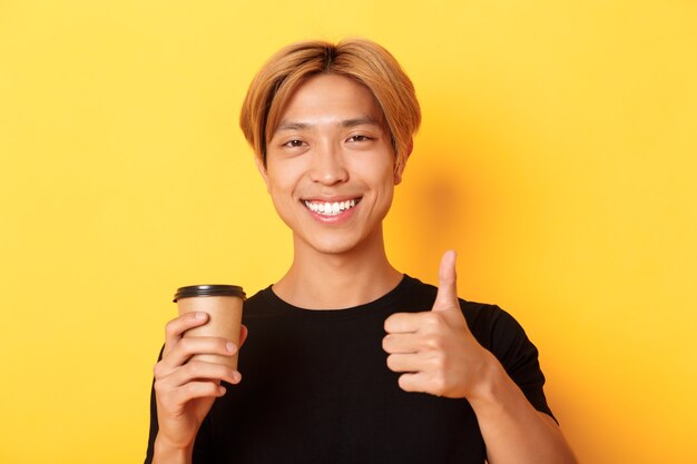 Primer plano de chico guapo asiático satisfecho recomendar café, sosteniendo una taza de café y mostrando el pulgar hacia arriba en señal de aprobación, sonriendo complacido sobre la pared amarilla