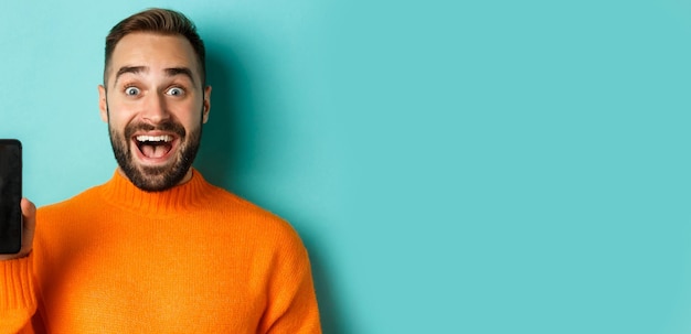 Foto gratuita primer plano de un chico guapo con barba en un suéter naranja que muestra la pantalla del teléfono inteligente y una sonrisa que muestra el baile de graduación