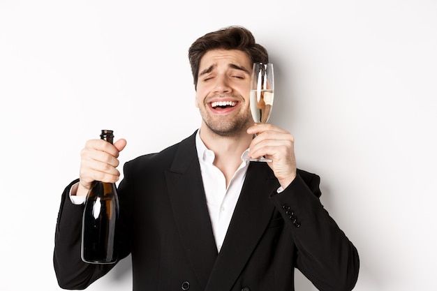 Primer plano de chico guapo borracho en traje, sosteniendo una copa de champán y celebrando el año nuevo, de pie sobre fondo blanco