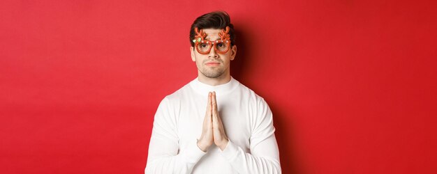 Primer plano de chico lindo en gafas de fiesta de Navidad, pidiendo ayuda, necesita un favor, de pie sobre fondo rojo.