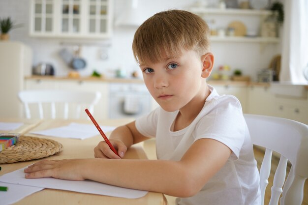 Primer plano de un colegial europeo en camiseta blanca sentado en la mesa de madera dibujando una imagen o haciendo los deberes con el interior de la cocina, mirando, con expresión facial seria