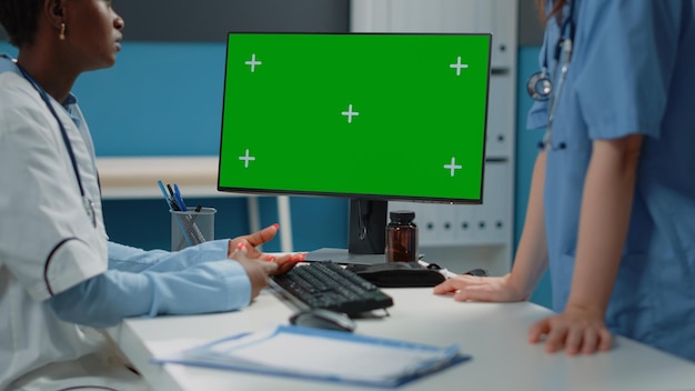 Primer plano de la computadora con pantalla verde horizontal en el escritorio mientras el médico y la enfermera hablan. Equipo médico usando monitor con plantilla de maqueta y fondo aislado para clave de croma.