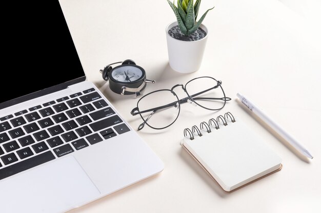 Primer plano de una computadora portátil abierta, gafas, una planta, un bolígrafo, un bloc de notas y un reloj despertador