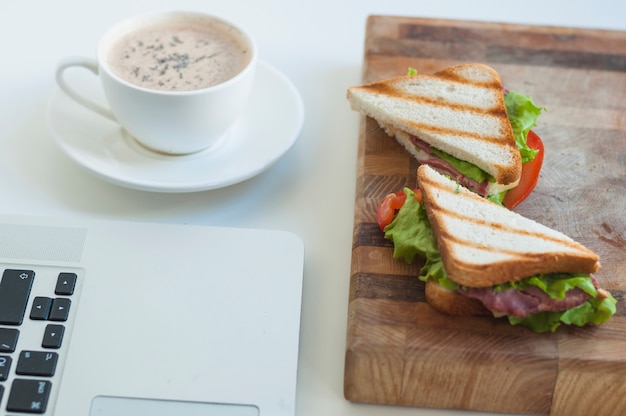 Primer plano de la computadora portátil; Taza de café y sándwiches en tabla de cortar contra el fondo blanco