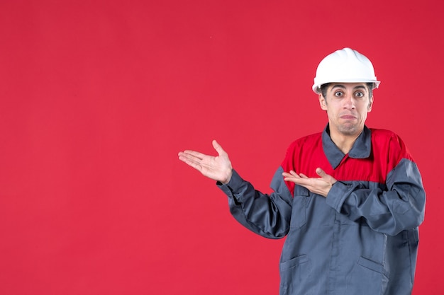 Primer plano de confundido joven trabajador en uniforme y con casco apuntando algo en el lado derecho en la pared roja aislada