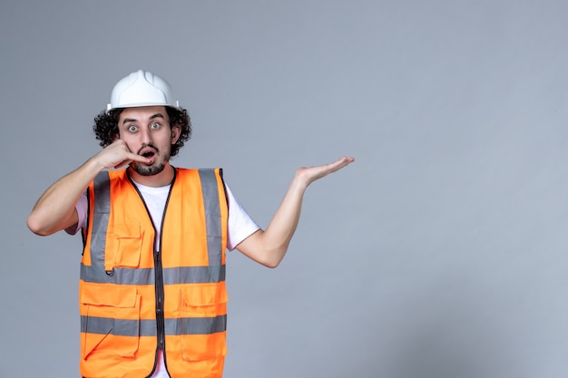 Primer plano de un constructor masculino conmocionado en un chaleco de advertencia con casco de seguridad y apuntando algo en el lado izquierdo y haciendo un gesto de llamarme sobre la pared gris de la ola