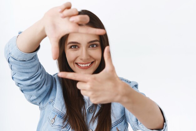 Primer plano creativo joven fotógrafa en busca de inspiración, mirando a través de una lente falsa, haciendo marcos con los dedos y sonriendo mientras se encuentra una increíble escena de disparo, fondo blanco