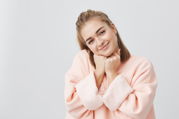 Primer plano de una curiosa mujer caucásica bonita con el pelo lacio y rubio, vestido con una cara de almohada rosa en sus manos y mirando suavemente, sonriendo mientras escucha historias interesantes