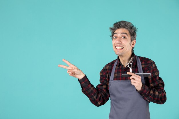 Primer plano de curioso joven barbero vistiendo delantal gris y sosteniendo el peine y la tijera mostrando dos sobre fondo de color azul