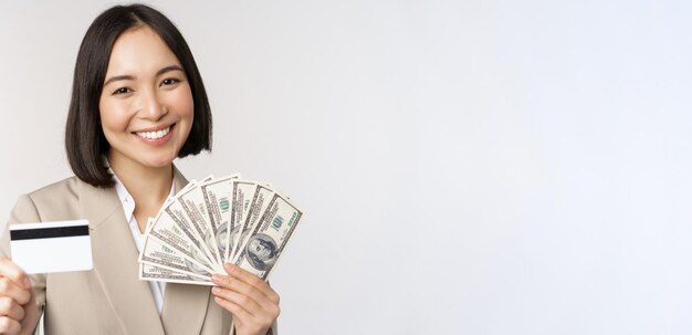 Primer plano de la dama de la oficina de la mujer de negocios asiática que muestra la tarjeta de crédito y los dólares de dinero de pie en traje sobre fondo blanco