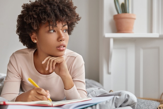 Primer plano de una dama pensativa de piel oscura con corte de pelo afro, mantiene la mano debajo de la barbilla, anota el texto en el bloc de notas con un bolígrafo amarillo, viste ropa informal, se acuesta en una cama cómoda, enfocado a un lado. Concepto de descanso