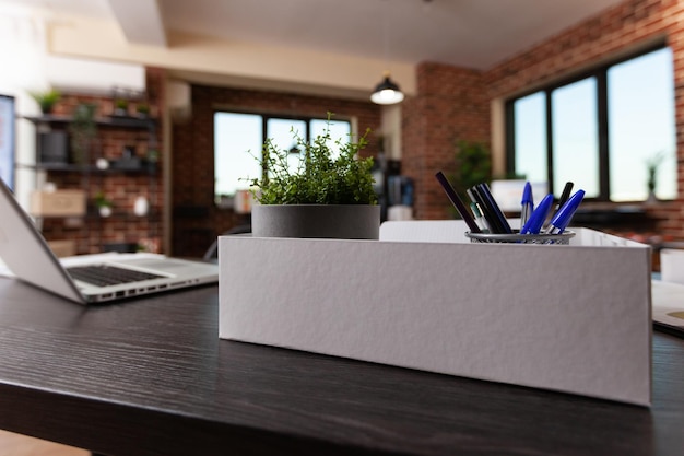 Primer plano de las decoraciones de oficina en la caja y el portátil en el escritorio. Planta de interior en maceta, suministros e instrumentos sobre una mesa de madera en un espacio comercial vacío. Maceta y adornos modernos en el trabajo.