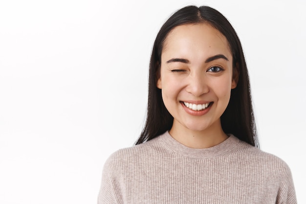 Primer plano descarado, entusiasta feliz sonriente mujer asiática con cabello largo y oscuro, maquillaje natural desnudo, tener un gran día, enviar vibraciones positivas, guiñar un ojo para animar a un amigo y decir todo bajo control