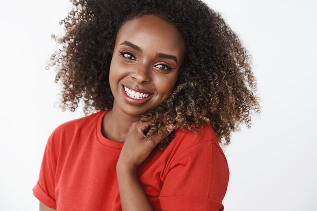 Primer plano de despreocupada, feliz y gentil hermosa modelo afroamericana con rizos sonriendo ampliamente tocando el cabello, mirando encantada, tierna al frente con camiseta roja sobre pared blanca