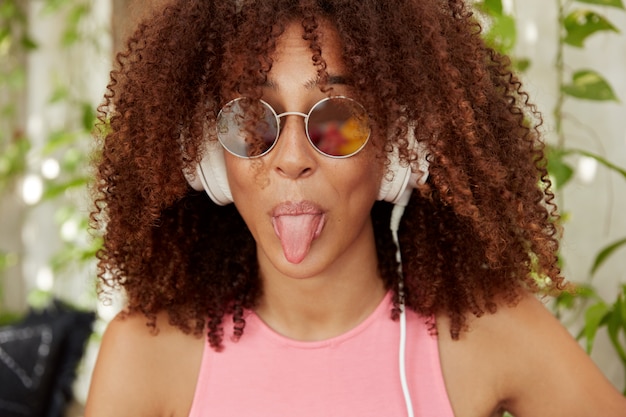 Foto gratuita primer plano de divertida modelo de mujer linda de piel oscura lleva gafas de sol, tiene el pelo rizado, escucha música en auriculares, muestra la lengua. bastante cómico joven afroamericana disfruta de una canción popular